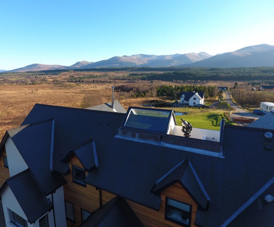 Aerial view of a modern house with a dark roof, situated in a mountainous landscape. Rolling hills and a vast field stretch in the background under a clear blue sky. Other houses are visible in the area, surrounded by greenery.